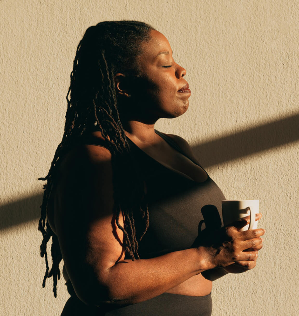 A woman holding a mug in both hands, eyes closed, in low sunlight against a warm wall