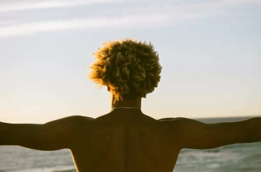 A person seen from behind, arms open to the sea at low sun