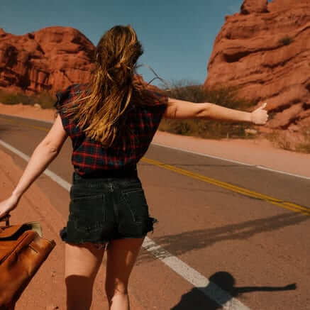 A woman walking a desert road with her thumb out, red rock behind her
