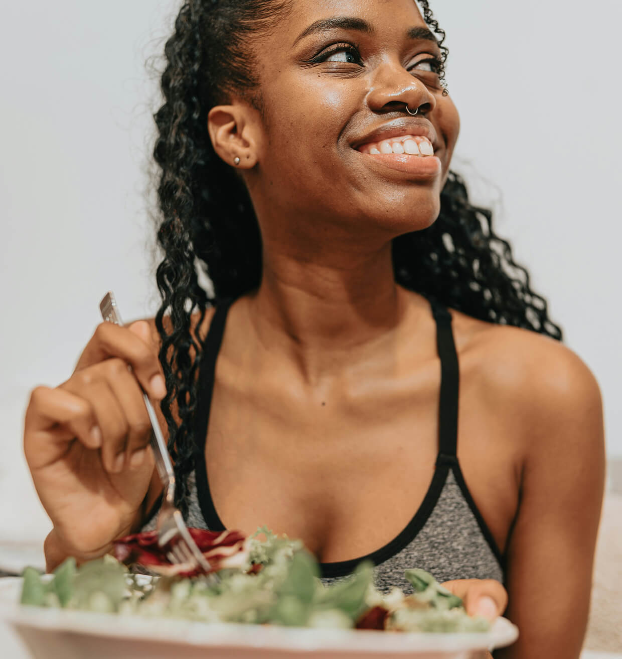 A woman in a vest top smiling over a bowl of salad, fork in hand